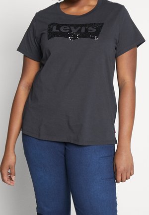 Frau trägt ein schwarzes Levi's T-Shirt mit kurzen Ärmeln und Pailletten-Logo sowie blaue Jeans, steht vor einem hellen Hintergrund.