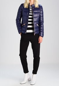 Marinblå puffjacka med kviltad design, dragkedjefickor och tryckknappsstängning, i kombination med en svart och vit randig tröja och svarta byxor. Vita stövlar.