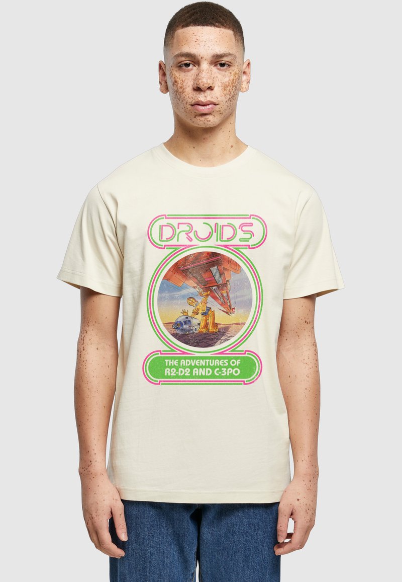 Jeune homme avec des taches de rousseur portant un T-shirt beige arborant les droïdes rétro Star Wars R2-D2 et C-3PO avec un texte et un graphisme néon.