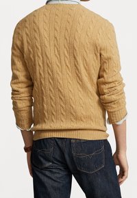 Pull en maille torsadée de couleur beige clair, avec un ourlet et des poignets côtelés. Porté sur une chemise rayée bleue et blanche, associé à un jean foncé.