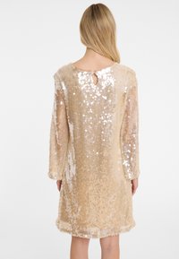 Robe en sequins dorés avec des manches longues, coupe fluide, encolure ronde et détail en trou de serrure au dos. Texture scintillante avec une doublure intérieure lisse.