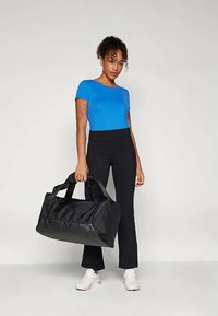Bolsa de deporte negra con asas dobles, fabricada en tejido duradero. Modelo vistiendo una camiseta azul y leggins negros, de pie con la bolsa sobre el hombro derecho.