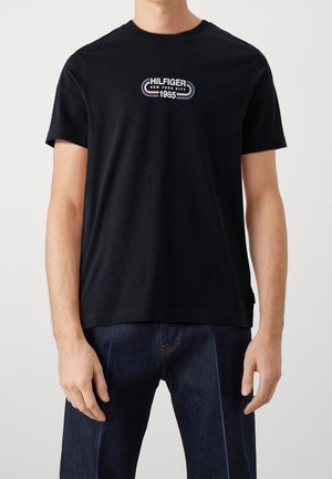 Man wearing a black short-sleeve T-shirt with "Hilfiger New York City 1985" logo and dark blue jeans, standing against a plain background.