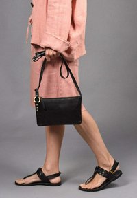 Clutch de cuero negro con cremallera, acento de anillo dorado y correa ajustable, combinado con un conjunto de lino rosa y sandalias negras.