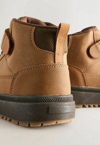 Braune synthetische Stiefel mit strukturiertem Material, verstärktem Knöchel, Zuglasche und Gummisohle mit tiefen Profilen für besseren Halt.