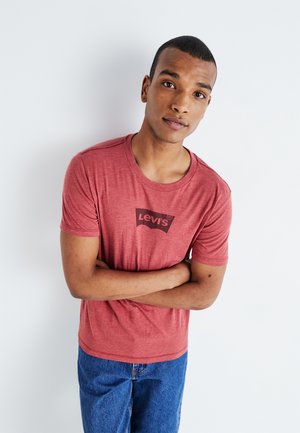 Camiseta de algodón roja con cuello redondo y mangas cortas, que presenta un logo negro de Levi's en la parte delantera. Se lleva con jeans de mezclilla azules.