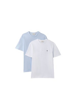 Mand iført en blå denimjakke over en hvid skjorte, lyse beige bukser, sort bælte og sorte sko, står med hænderne delvist i lommerne.