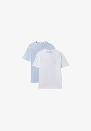 Homme portant une veste en jean bleue sur une chemise blanche, pantalon beige clair, ceinture noire et chaussures noires, debout avec les mains partiellement dans les poches.