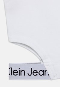 Wit katoenen T-shirt met een ronde hals, voorzien van een wit elastisch bandje met structuur aan de onderkant waarop "Klein Jeans" in zwart is gedrukt.