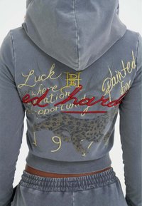Grijze cropped hoodie met capuchon, voorzien van geborduurde tekst in goud en rood, een luipaardprintontwerp en een zachte, gestructureerde stof.