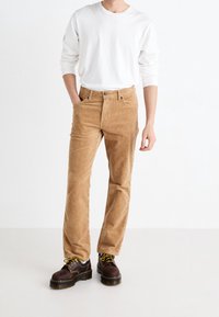 Světlé hnědé kordové kalhoty s rovým střihem, které mají přední kapsy a zapínání na knoflík. Nošeny s bílou košilí s dlouhým rukávem a hnědými botami.