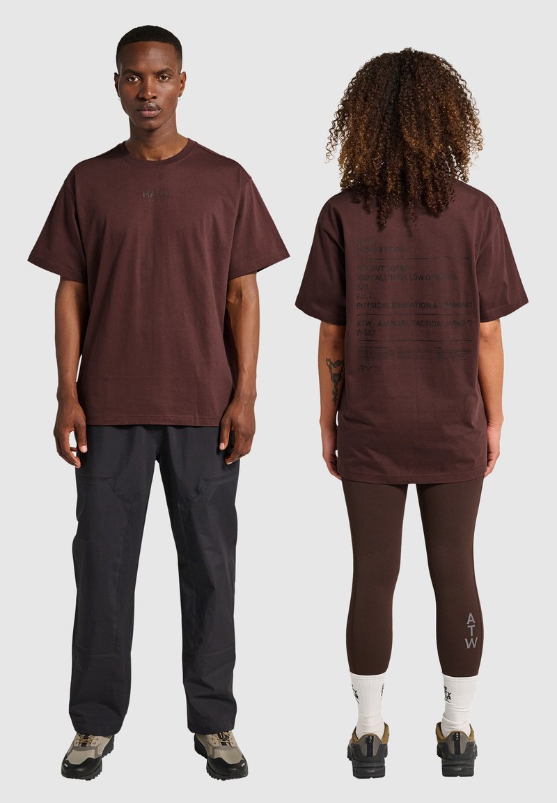 Brun bomullströja med korta ärmar, har tryckt text i svart på baksidan, baren med mörka byxor och bruna leggings.