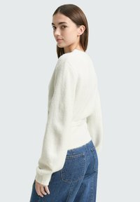 Bela pletena pulover z teksturiranim zaključkom in spušenimi rameni, z zategnjenim pasom in dolgimi rokavi s manšetami. Oblikovan z modrimi kavbojkami.