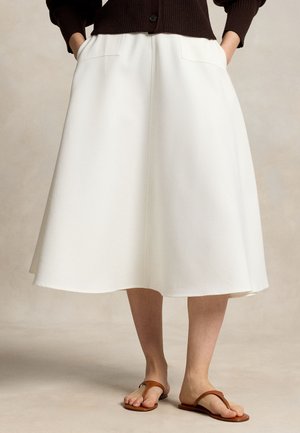 Femme portant une jupe midi blanche en ligne A avec des poches à l'avant, associée à un pull marron et des sandales en cuir marron, debout les mains dans les poches.