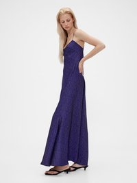 Langes lila Kleid mit dünnen Trägern, das ein Pünktchenmuster aufweist und aus einem fließenden, strukturierten Stoff besteht. Kombiniert mit schwarzen, flachen Sandalen.
