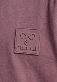 Tejido púrpura con un parche de goma que presenta el nombre de la marca "hummel" y un diseño estilizado, cosido al material.