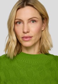 Hellgrüner Strickpullover mit strukturiertem Honigwabenmuster und einem gerippten Ausschnitt. Getragen von einer Person mit blondem Haar.