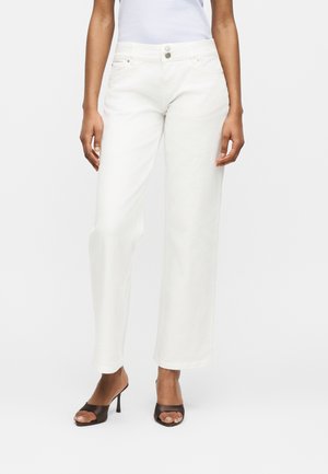 Bas du corps d'une personne portant un jean blanc à jambes larges et des sandales à talons noires ouvertes à l'avant, debout devant un fond uni.