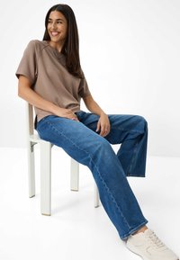 Bruine top met korte mouwen, blauwe wijde jeans en lichtgekleurde sneakers. Model zit op een witte stoel met gouden accenten.