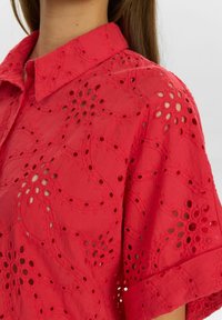 Blusa rossa in pizzo con colletto, modelli intagliati intricati e un tessuto leggero dall'aspetto texturizzato.