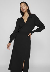 Juodas megztas suknelė su V formos iškirpte ir apykakle, ilgomis pūstomis rankovėmis ir šonine praskilimu. Sklandus audinys ir prigludusi prie liemens.