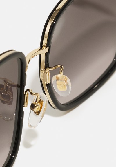 Black sunglasses with a thick frame, gold accents, and gradient gray lenses. Features adjustable nose pads with a logo detail.