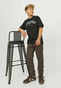 Čierne tričko s bielym logom "JACK & JONES", v kombinácii s hnedými cargo nohavicami a hnedými teniskami, stojí vedľa čierneho kovového stoličky.