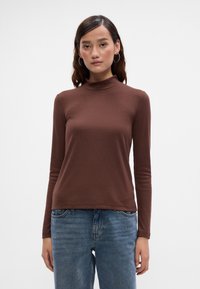 Egy nő testhez simuló barna bordás garbót és kék farmernadrágot visel, egy sima fehér háttér előtt áll.