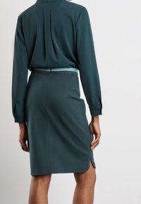 Kvinna i en teal långärmad blus som är stoppad i en matchande knälång kjol med en subtil slits på ena sidan.