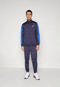 Fato de treino azul-marinho com detalhes em azul, feito de tecido macio. Casaco com fecho de correr e bolsos laterais, combinado com calças ajustadas. Ténis desportivos brancos.