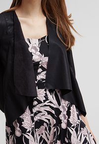 Svart konstläderjacka med öppen front och kraftig textur, lager över en blommönstrad klänning med rosa och vita blommor på svart bakgrund.