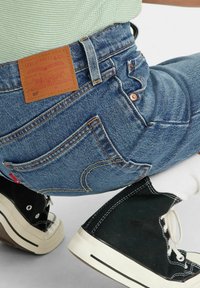 Jeans in denim blu con una toppa posteriore in pelle marrone, caratterizzati da cuciture arancioni e tasche. Abbinati a sneakers alte in tela nere.