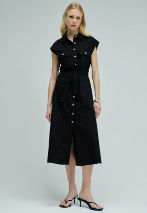 Mujer vistiendo un vestido midi negro con botones delanteros, mangas ranglán, cinturón en la cintura, bolsillos delanteros, pulsera plateada y sandalias negras de tacón.