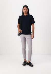 Marineblaues Polo-Shirt mit strukturiertem Design, kombiniert mit hellgrauen Slim-Fit-Hosen und schwarzen Lederloafern, Model hält eine Tasche.