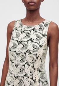 Gros plan d'une robe sans manches avec des motifs de feuilles en dentelle crème sur un tissu noir portée par une personne à la peau foncée.