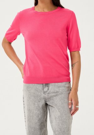 Vrouw draagt een felroze gebreide top met korte mouwen in combinatie met lichtgrijze gewassen jeans, met een tatoeage op de linkeronderarm en zilveren ringen aan de rechterhand.