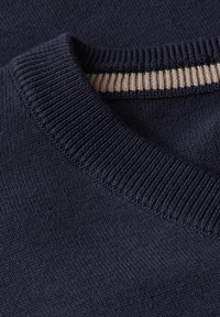 Mörkblått stickat tröja med en strukturerad, ribbad krage som har en kontrasterande beige rand. Närbild visar tygdetaljer.