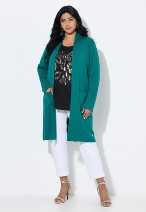 Cardigan largo en teal de tela suave con apertura frontal, dos bolsillos y un botón de acento dorado. Lleva puesto sobre una blusa negra con lentejuelas y pantalones blancos.