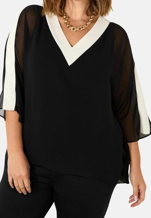 Vrouw draagt een zwarte V-hals blouse met witte bies en doorzichtige mouwen, gecombineerd met een zwarte broek en een dikke gouden ketting.