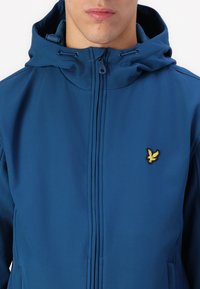 Blå zipjacka med huva, prydd med en gul broderad logotyp på vänster bröst. Slätt material och skräddarsydd passform med framfickor.