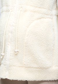 Κοντινό πλάνο κρεμ Sherpa fleece μπουφάν με φερμουάρ, μπροστινή τσέπη και κορδόνια με χάντρες.