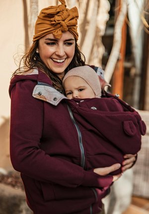 Bordeauxkleurige hoodie met een rits aan de voorkant, zijzakken en een afneembare babydraagfunctie. Zacht materiaal en een gewatteerde voering zijn zichtbaar.