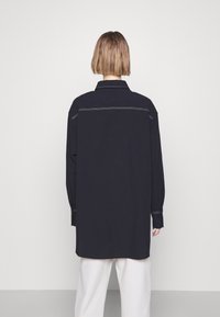 Chemise bleu marine à manches longues avec col, présentant des détails de coutures blanches, une coupe droite et des poignets boutonnés. Portée par-dessus un pantalon de couleur claire.