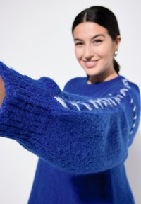 Kongelig blå strikket sweater med tykke ribbede ærmer, med hvide detaljer langs skuldersømmen. Blød tekstur med en løs pasform.