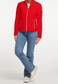 Červená péřová bunda s vysokým límcem a vodorovným prošíváním. Má přední zip a dvě boční kapsy. Kombinováno s modrými džíny s rovnými nohavicemi a bílými teniskami.