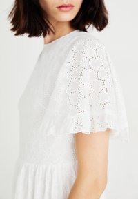 Witte katoenen jurk met hartpatroon in broderie anglaise, korte geruite mouwen en een aansluitend lijfje met een uitlopende rok.