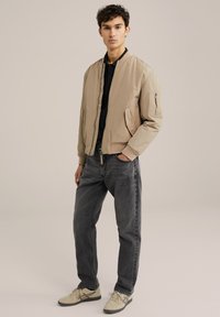 Beige bomber jacket witha zip closure, two side pockets, and a pocket on the left sleeve. Worn over a black polo shirt and paired with dark jeans and sneakers.