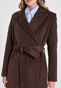 Cappotto avvolgente marrone, strutturato, con cintura in vita, ampi revers e tasche laterali. Indossato sopra una camicia azzurra con righe.