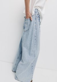 Lichtblauwe wijde denim jeans met een hoge taille, voorzien van zijzakken en een vervaagd uiterlijk, getoond met een hand in de zak.