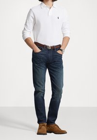 Bílá polokošile s dlouhým rukávem a límečkem, tmavě modré džíny, hnědý kožený opasek a hnědé semišové boty. Model stojí s rukama v kapsách.